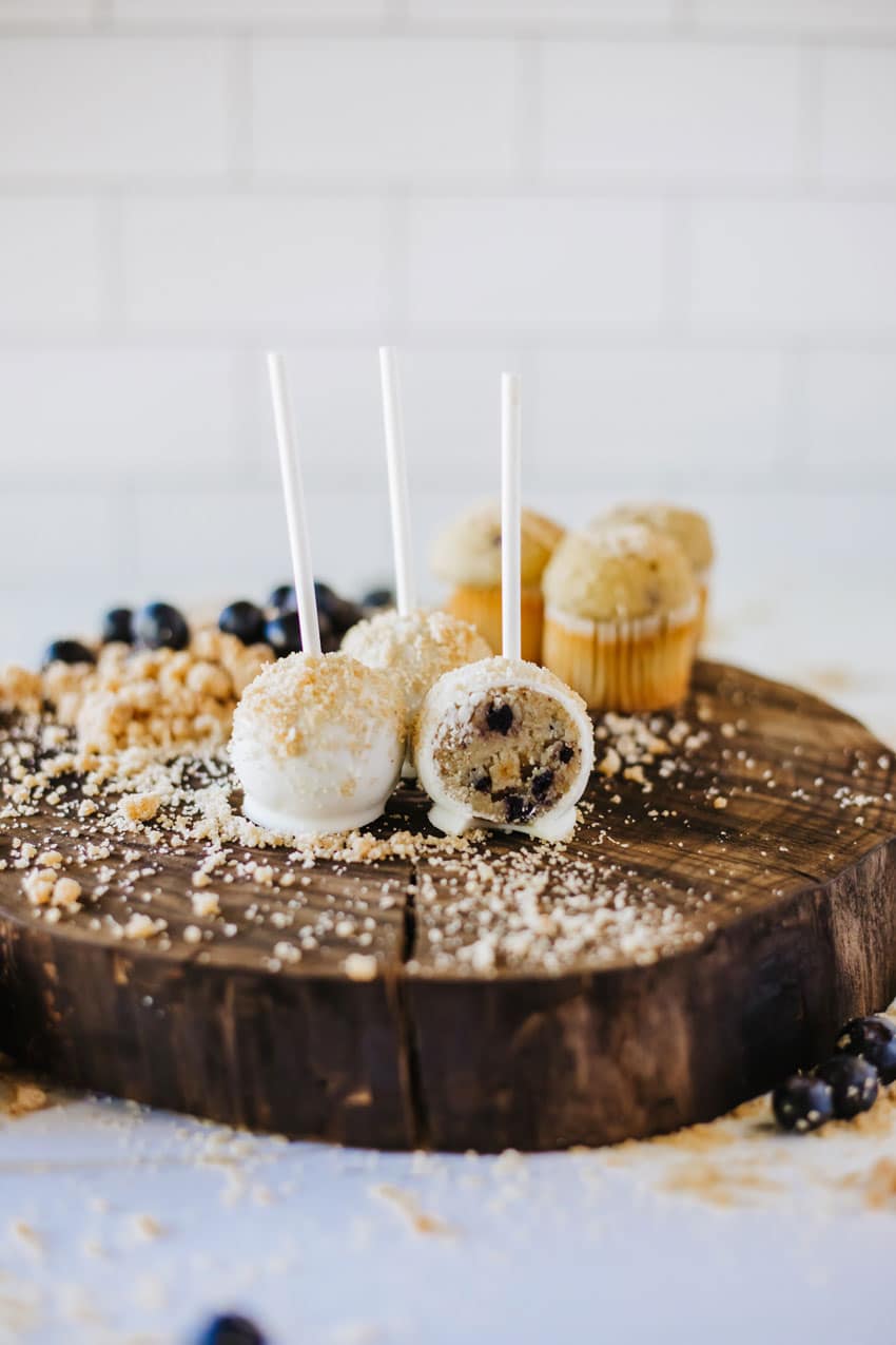 two halves of a blueberry muffin cake pop on a wooden table surrounded by graham cracker crumbs and fresh blueberries