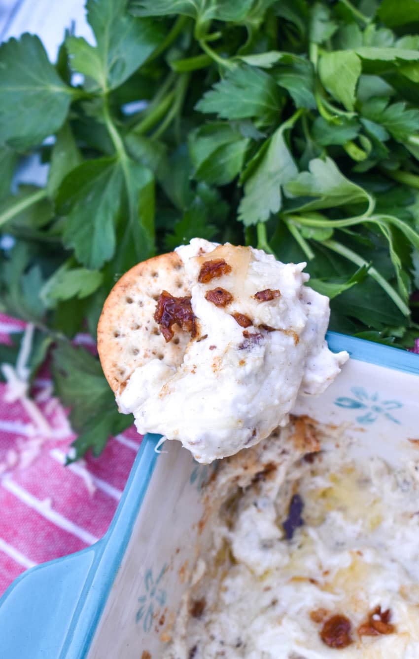 a cracker topped with a scoop of creamy caesar dip with bacon resting on the corner of a blue baking dish