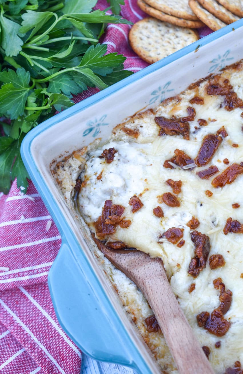 a wooden spoon scooping creamy caesar dip with bacon out of a small blue baking dish