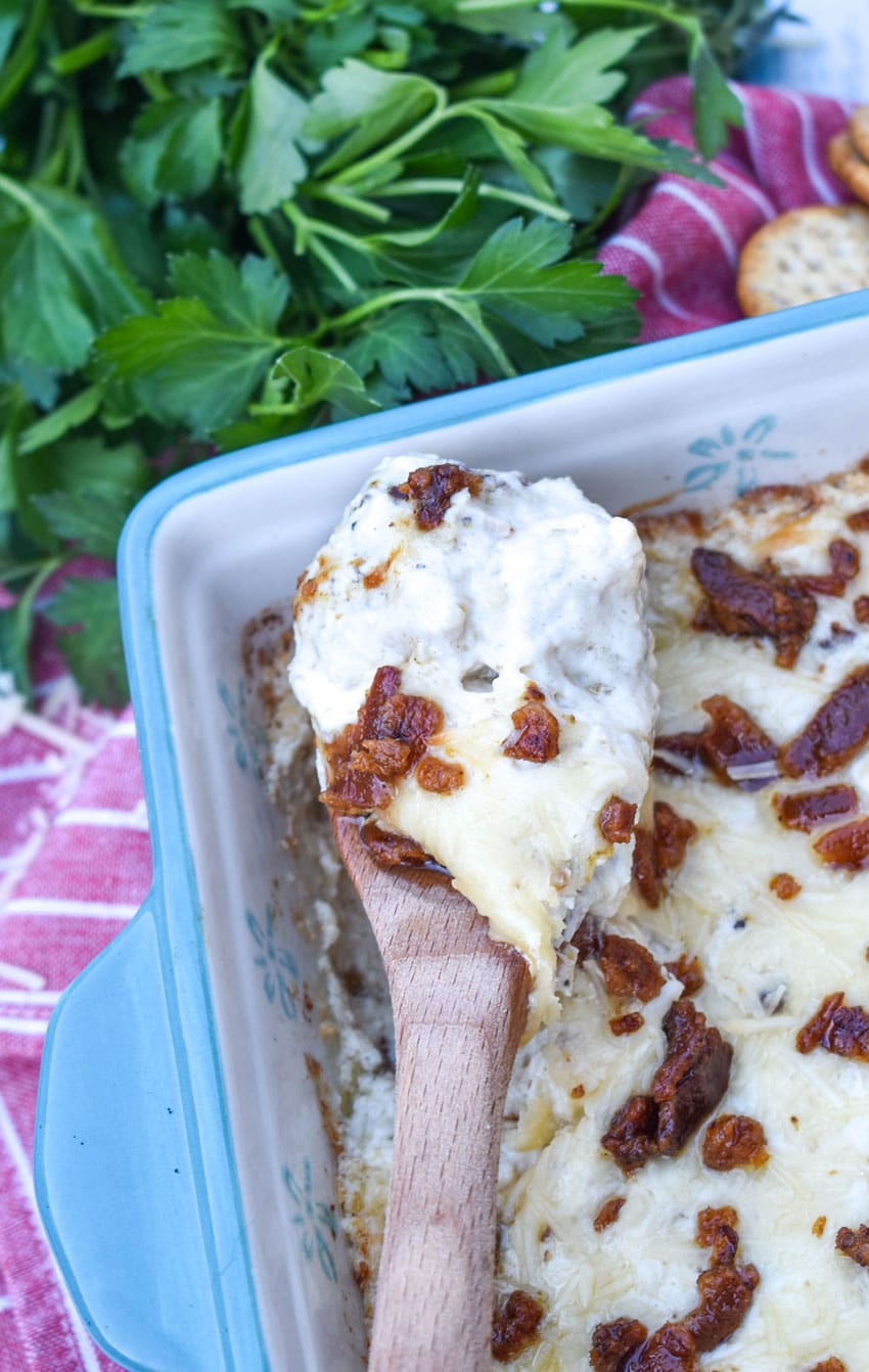 a wooden spoon holding up a scoop of creamy caesar dip with bacon