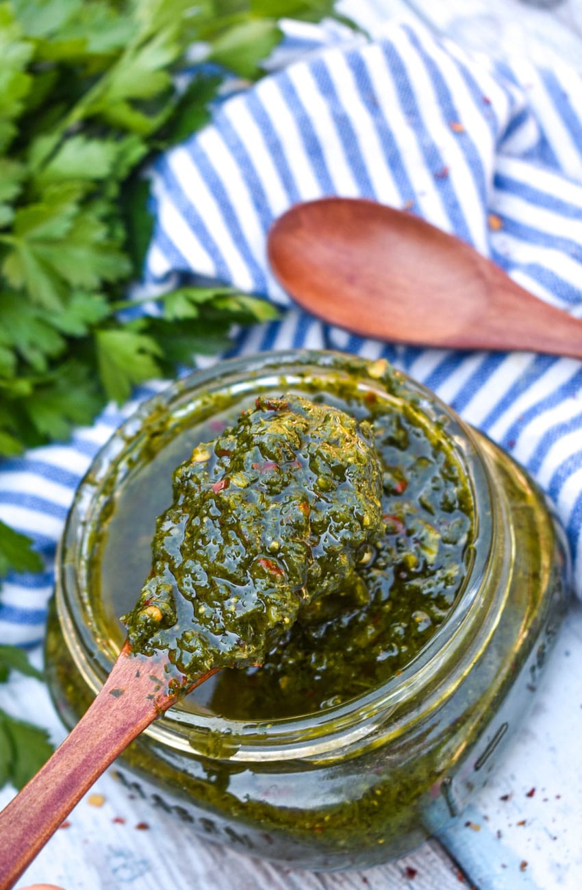 A WOODEN SPOON SCOOPING HOMEMADE CHIMICHURRI SAUCE OUT OF A SMALL GLASS JAR