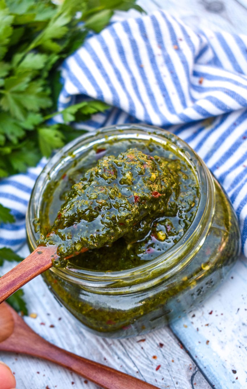 A WOODEN SPOON SCOOPING HOMEMADE CHIMICHURRI SAUCE OUT OF A SMALL GLASS JAR