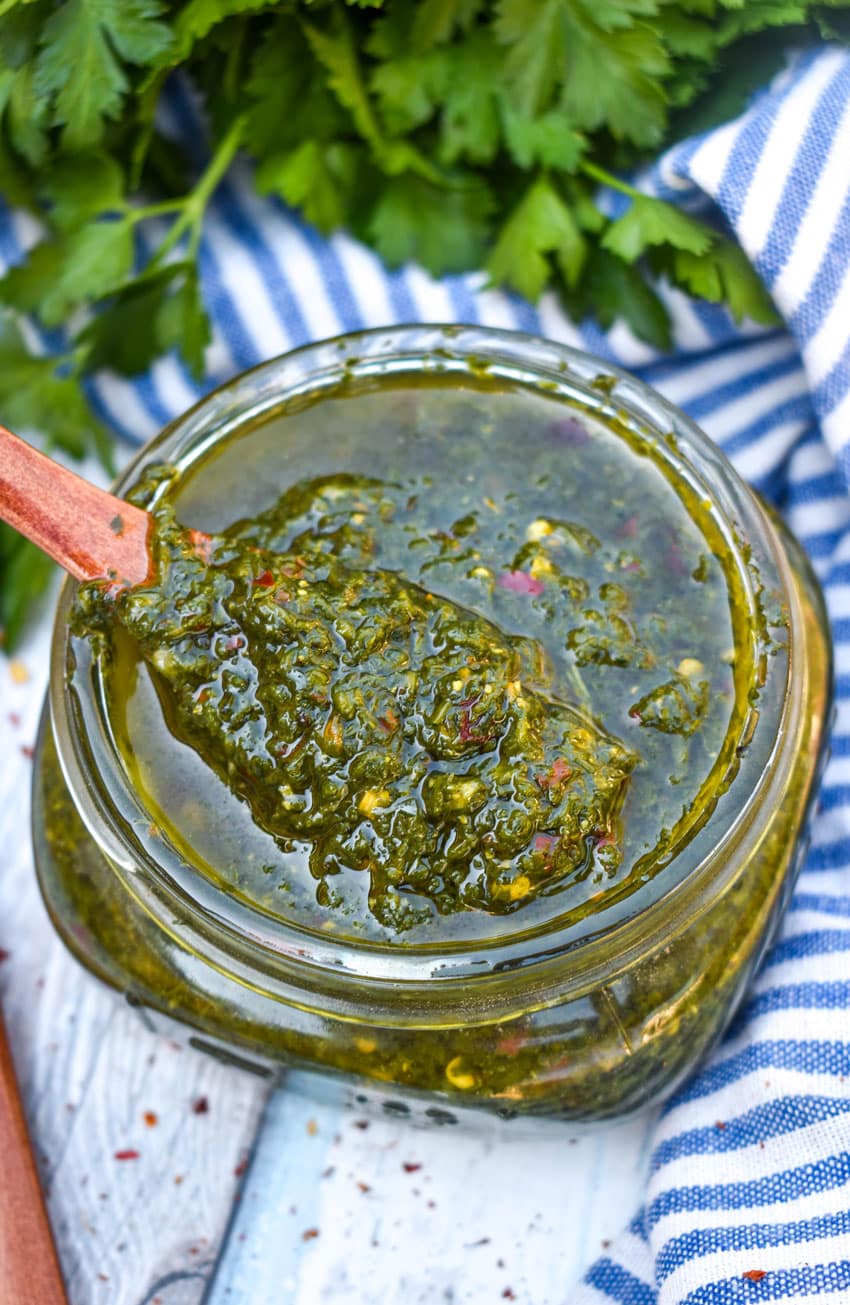 A WOODEN SPOON SCOOPING HOMEMADE CHIMICHURRI SAUCE OUT OF A SMALL GLASS JAR