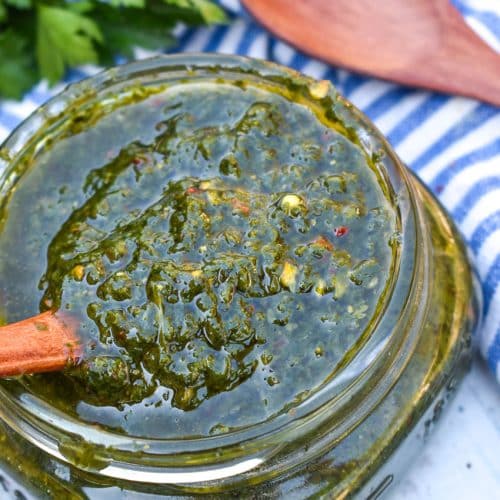 A WOODEN SPOON SCOOPING HOMEMADE CHIMICHURRI SAUCE OUT OF A SMALL GLASS JAR