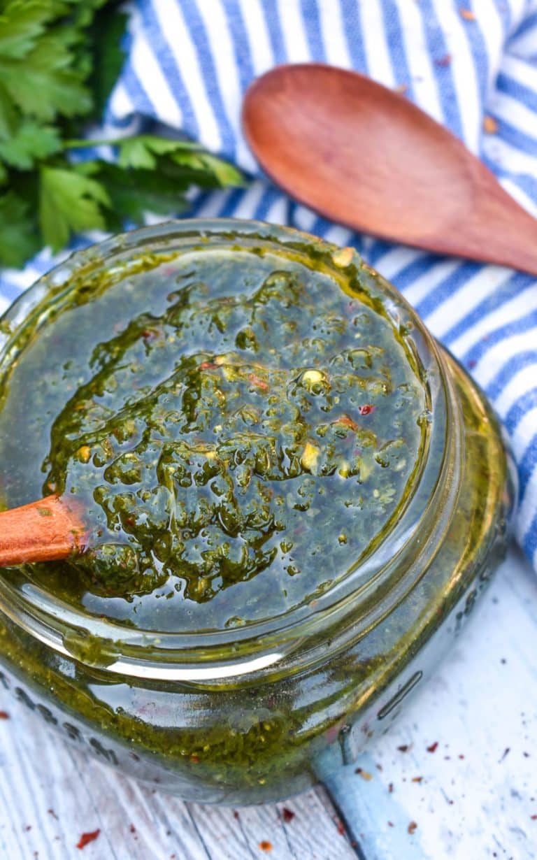 A WOODEN SPOON SCOOPING HOMEMADE CHIMICHURRI SAUCE OUT OF A SMALL GLASS JAR