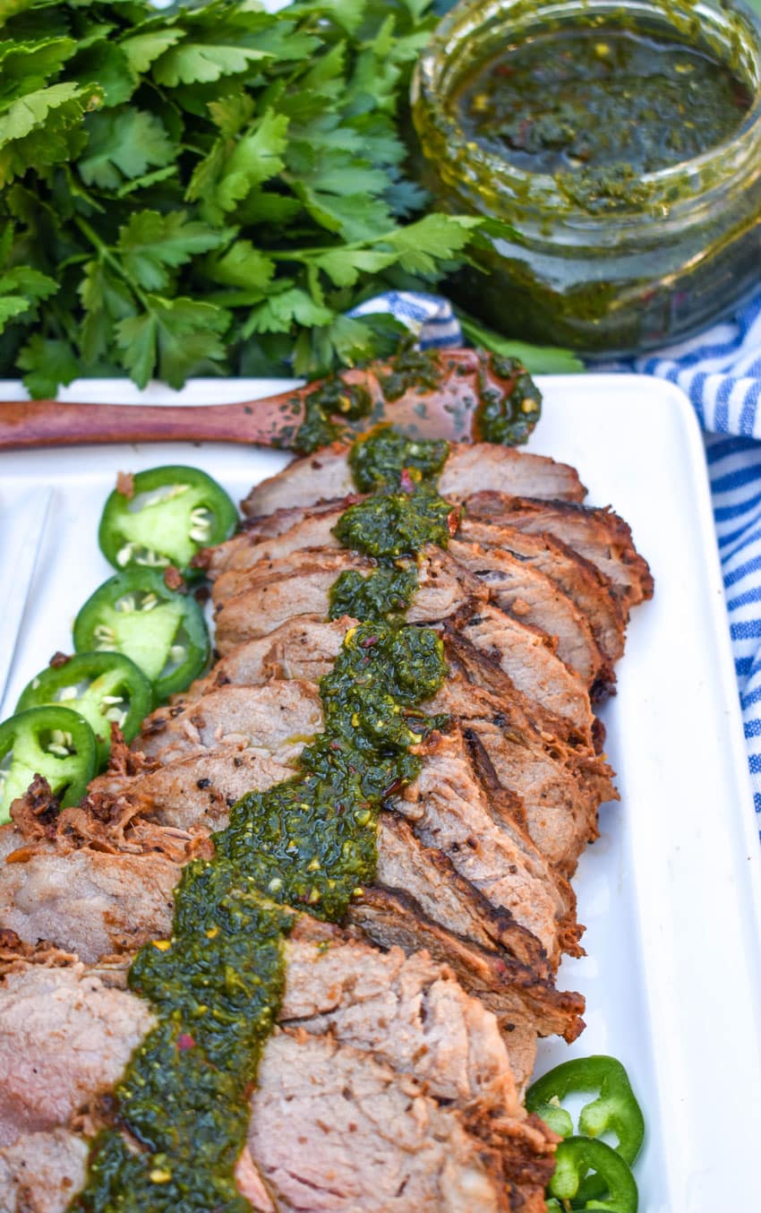 slices of smoked tri tip topped with homemade chimichurri sauce arranged in a row on a white serving platter