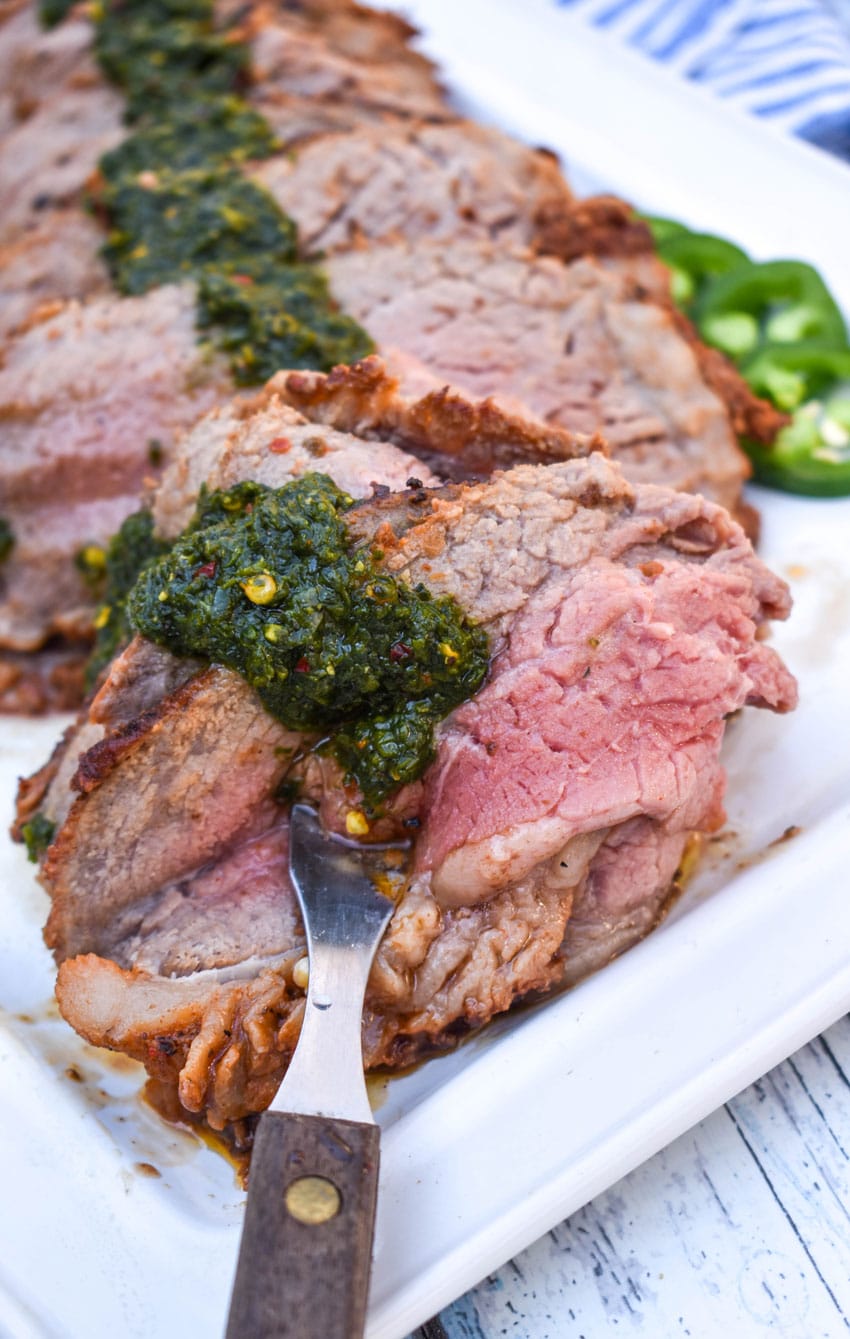 a meat fork scooping slices of smoked tri tip topped with homemade chimichurri sauce off a white platter