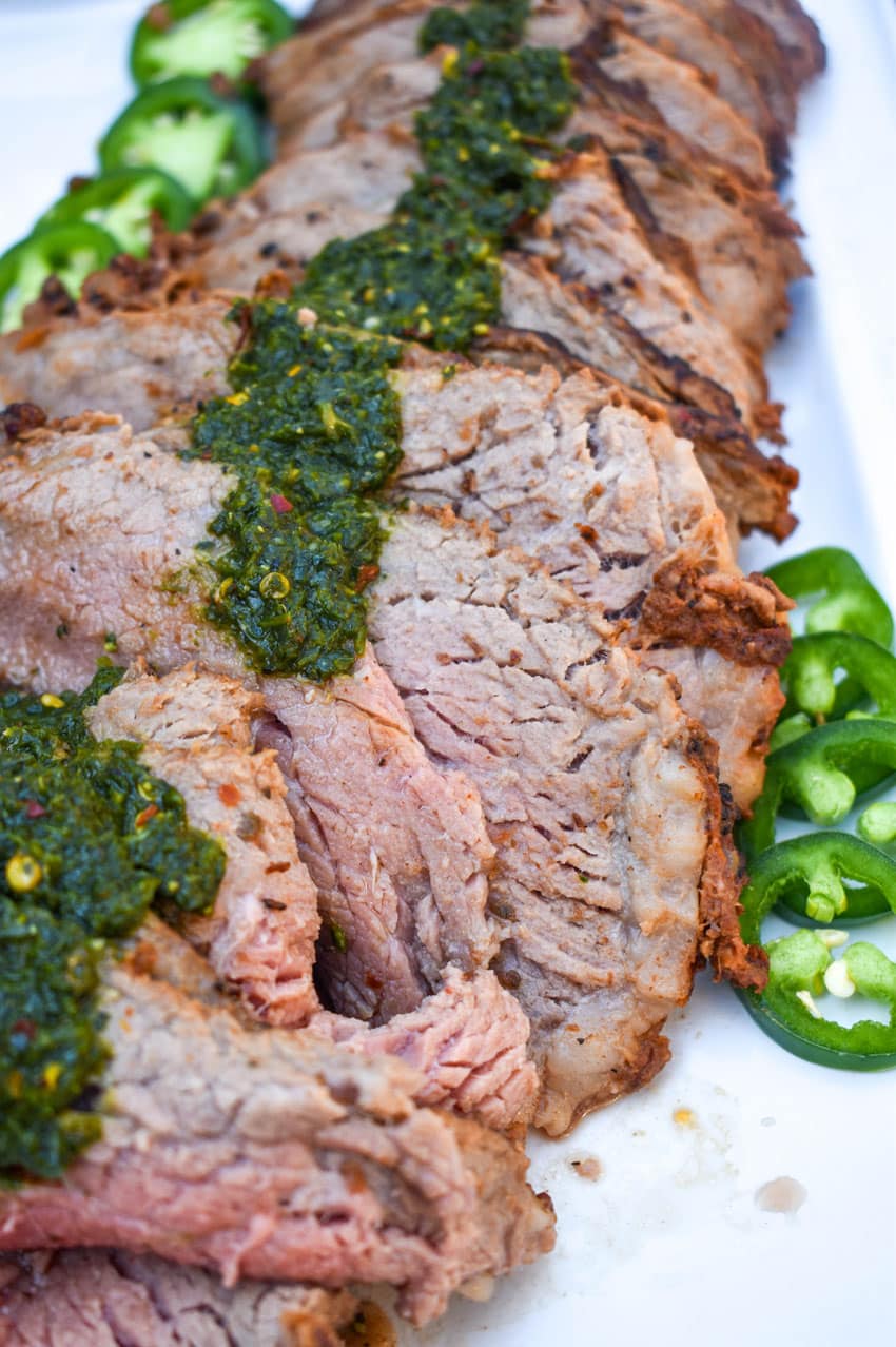 slices of smoked tri tip topped with homemade chimichurri sauce arranged in a row on a white serving platter