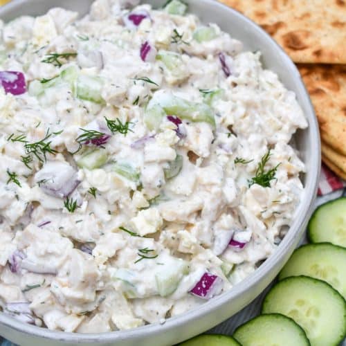 tzatziki chicken salad in a gray bowl surrounded by sliced cucumber, sliced lemons, and slices of fresh pita bread