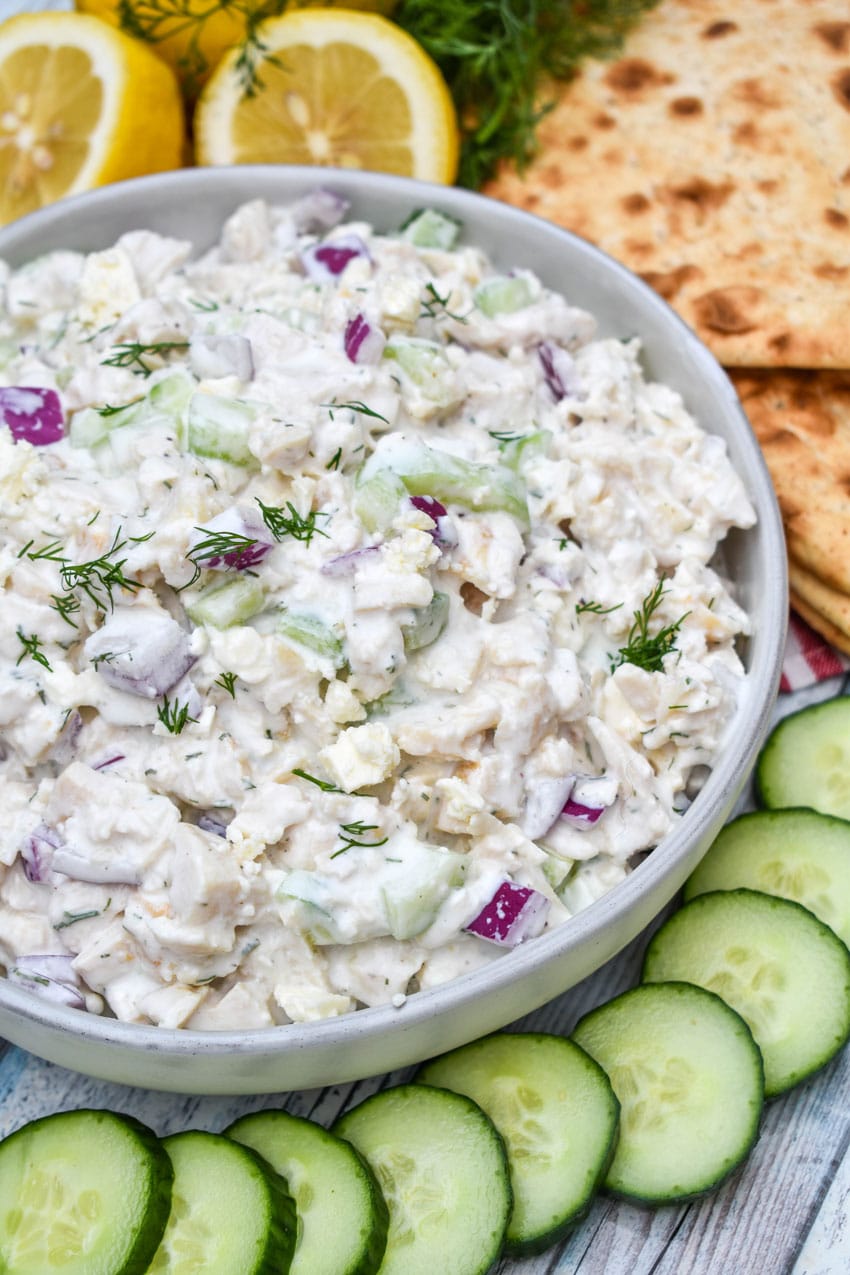 tzatziki chicken salad in a gray bowl surrounded by sliced cucumber, sliced lemons, and slices of fresh pita bread