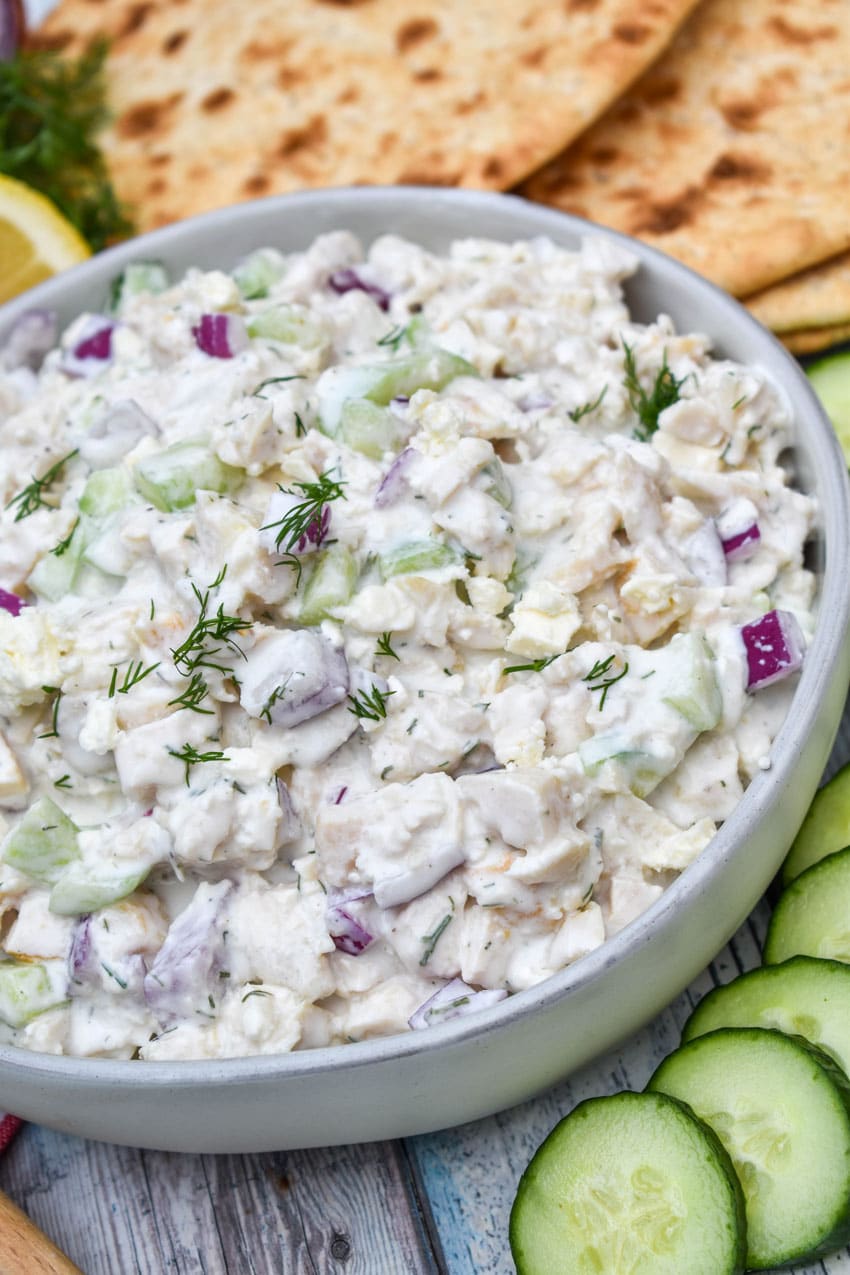 tzatziki chicken salad in a gray bowl surrounded by sliced cucumber, sliced lemons, and slices of fresh pita bread