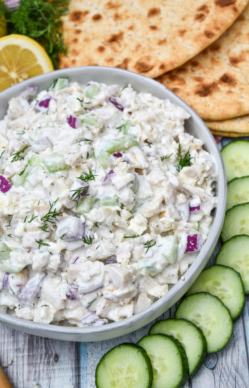 tzatziki chicken salad in a gray bowl surrounded by sliced cucumber, sliced lemons, and slices of fresh pita bread