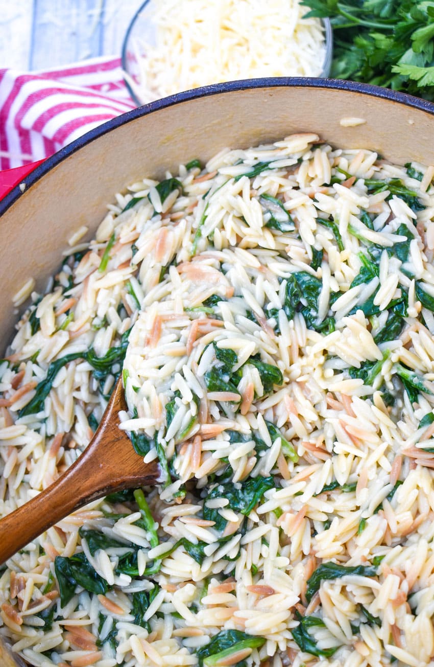 a wooden spoon scooping spinach parmesan orzo out of a red dutch oven