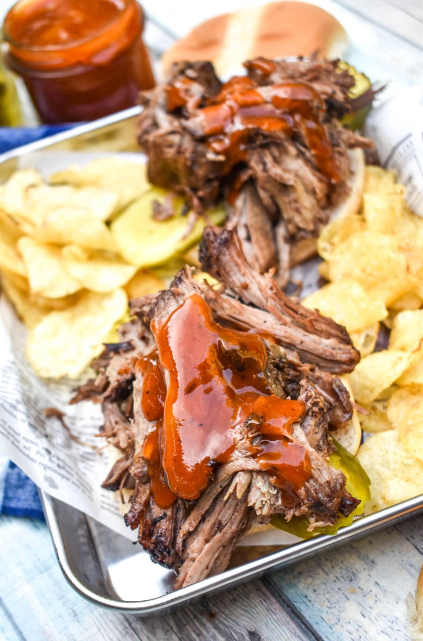 smoked chuck roast topped with barbecue sauce on a hamburger bun surrounded by potato chips on a parchment lined metal serving tray