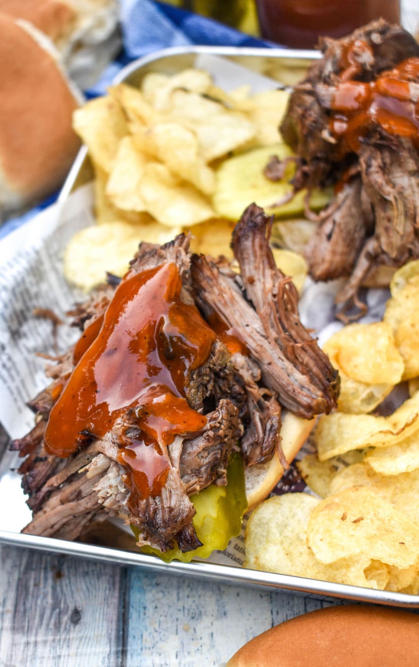 smoked chuck roast topped with barbecue sauce on a hamburger bun surrounded by potato chips on a parchment lined metal serving tray