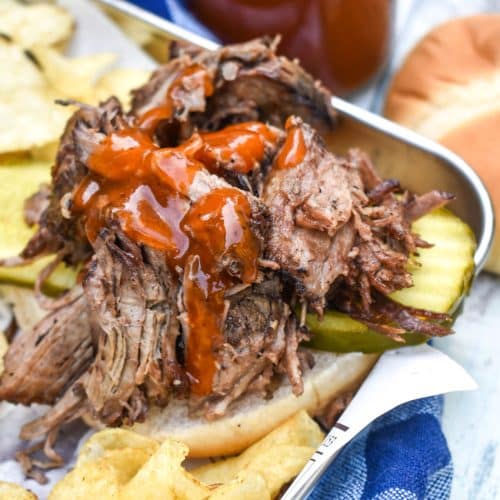 smoked chuck roast topped with barbecue sauce on a hamburger bun surrounded by potato chips on a parchment lined metal serving tray