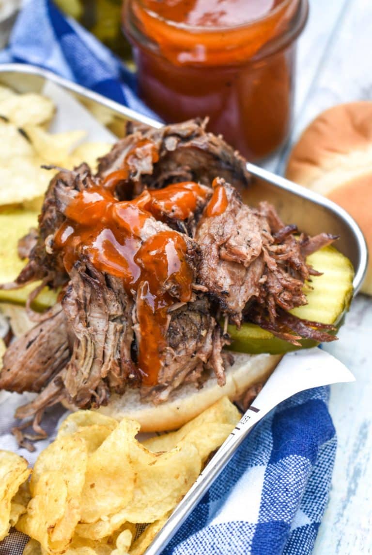 smoked chuck roast topped with barbecue sauce on a hamburger bun surrounded by potato chips on a parchment lined metal serving tray