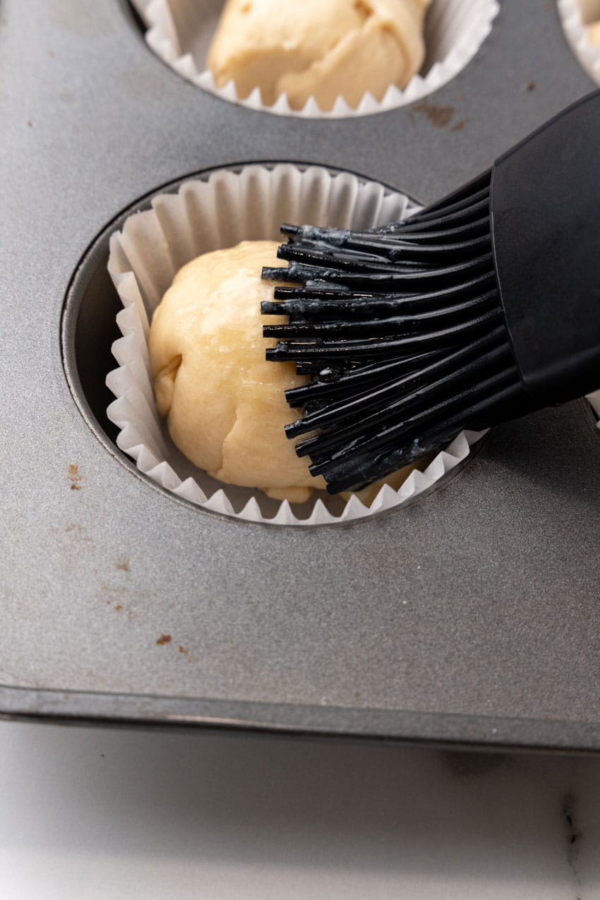 a pastry brush rubbing melted butter over an unbaked resurrection roll in a metal muffin pan