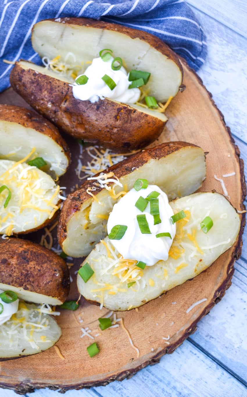 smoked baked potatoes topped with shredded cheese, sour cream, and sliced green onions on a wooden cutting board