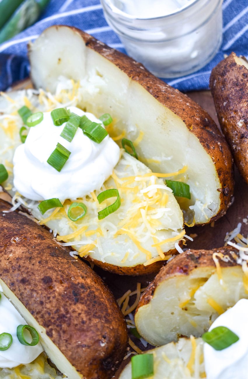 smoked baked potatoes topped with shredded cheese, sour cream, and sliced green onions on a wooden cutting board