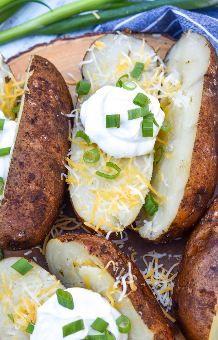 smoked baked potatoes topped with shredded cheese, sour cream, and sliced green onions on a wooden cutting board