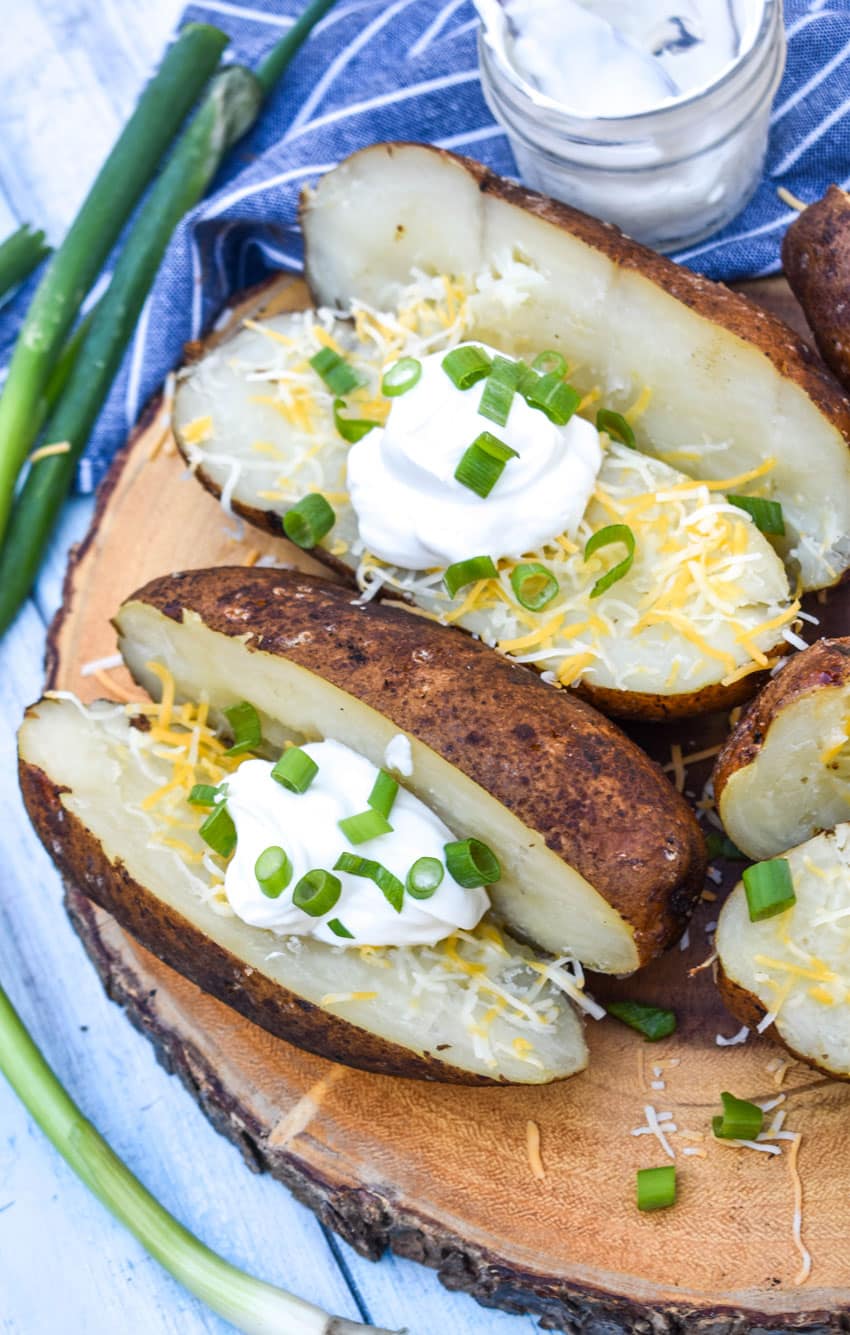 smoked baked potatoes topped with shredded cheese, sour cream, and sliced green onions on a wooden cutting board