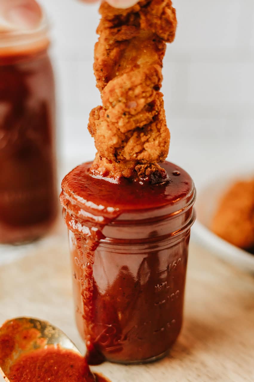 a crispy chicken tender being dipping in a jar of homemade barbecue sauce