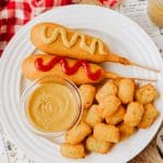 homemade yellow mustard in a small glass jar on a white plate next to corn dogs and tater tots