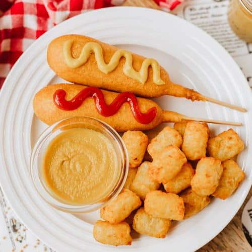 homemade yellow mustard in a small glass jar on a white plate next to corn dogs and tater tots