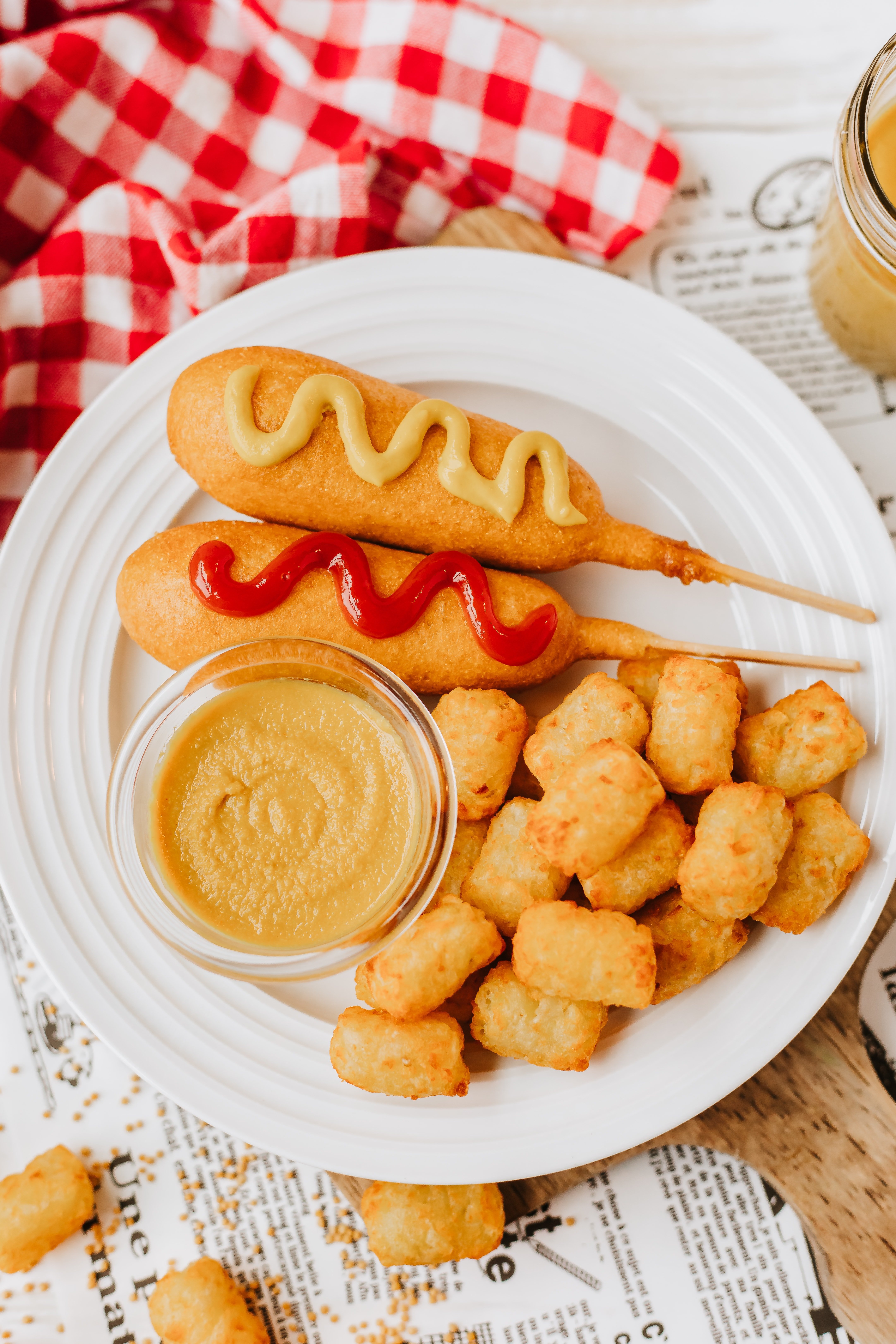 homemade yellow mustard in a small glass jar on a white plate next to corn dogs and tater tots