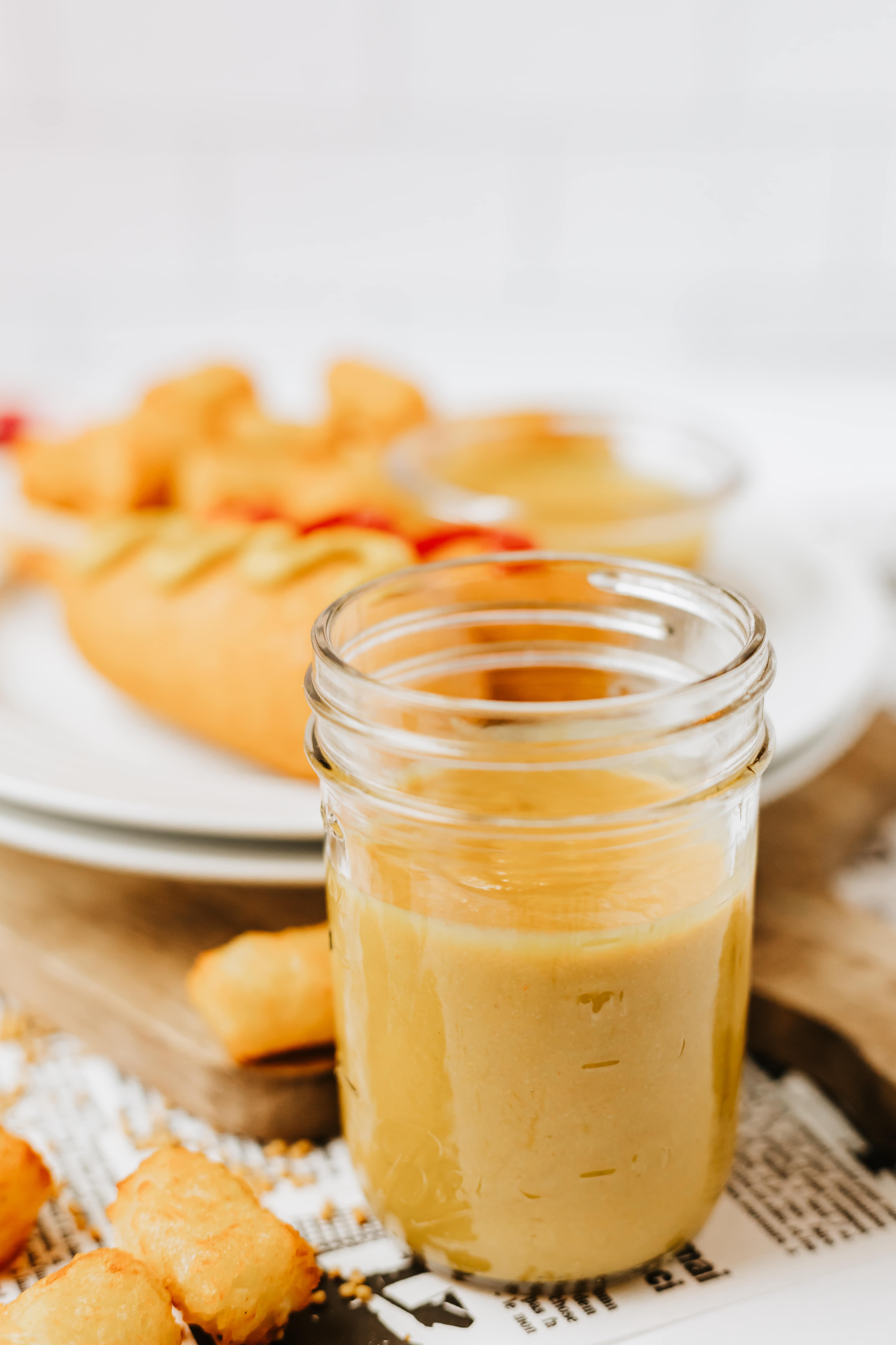 homemade yellow mustard in a small glass jar on a white plate next to corn dogs and tater tots