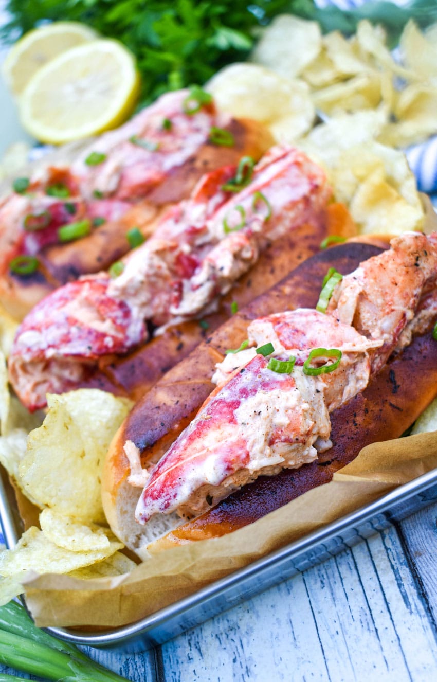 three homemade lobster rolls on a metal serving pan with potato chips on the sides
