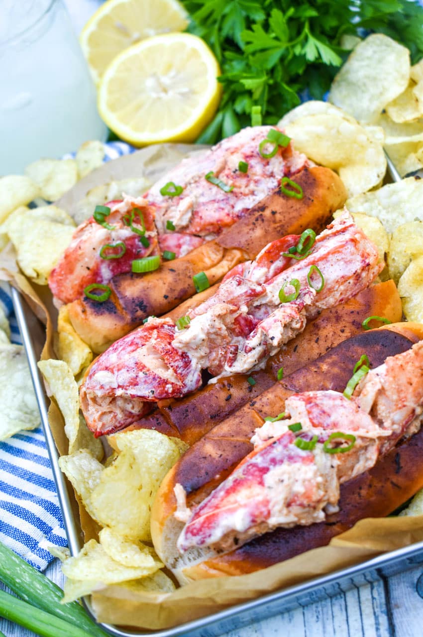 three homemade lobster rolls on a metal serving pan with potato chips on the sides