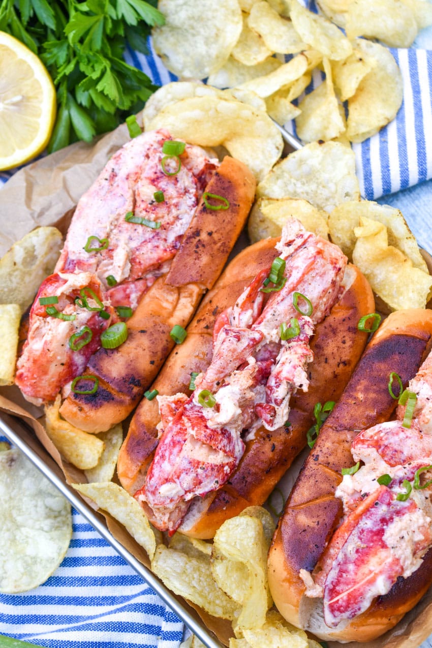 three homemade lobster rolls on a metal serving pan with potato chips on the sides
