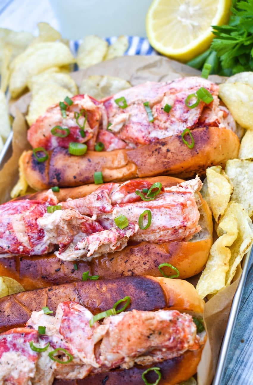 three homemade lobster rolls on a metal serving pan with potato chips on the sides