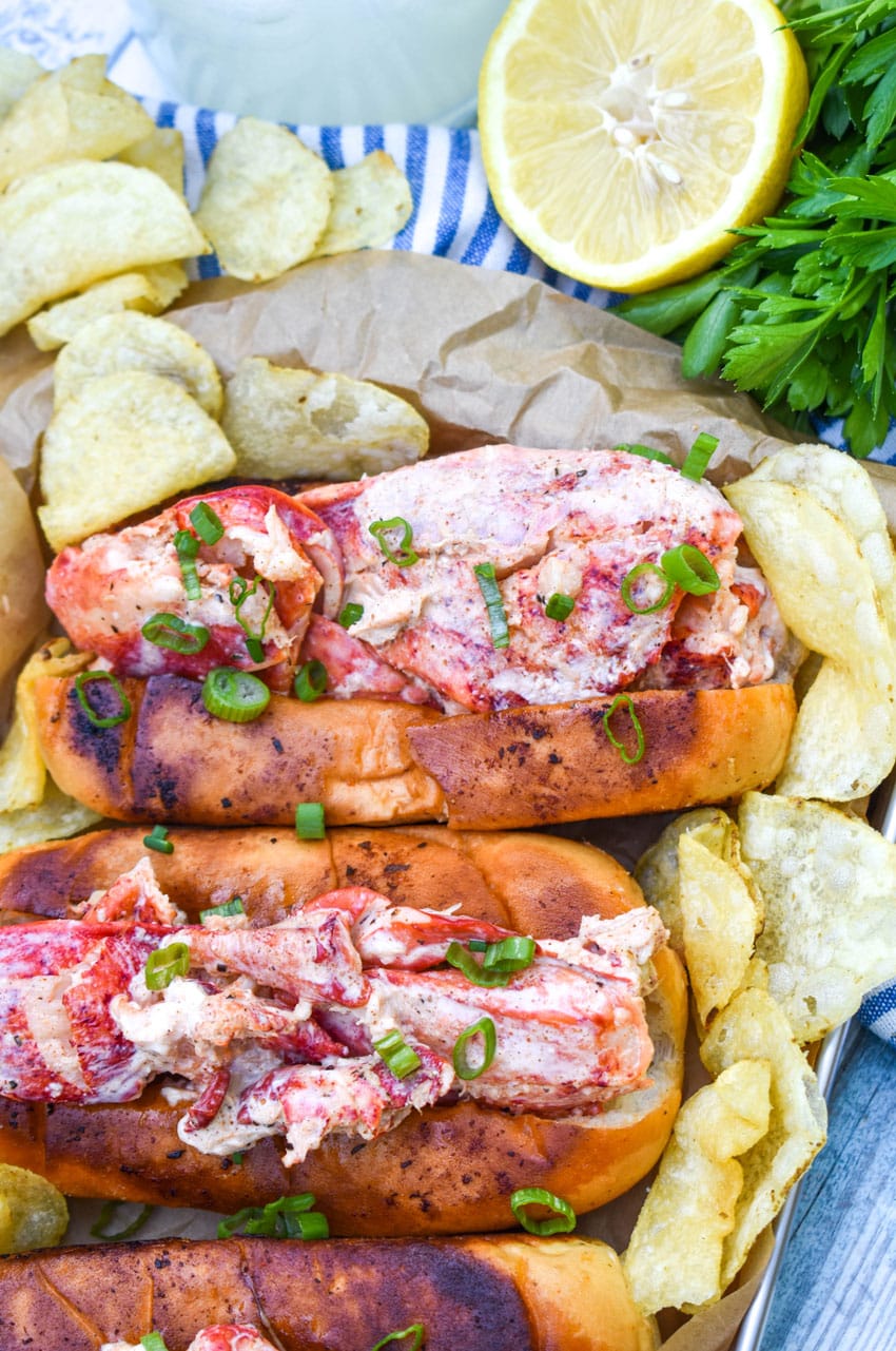 three homemade lobster rolls on a metal serving pan with potato chips on the sides