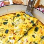 a wood handled spatula lifting a slice of garden vegetable fritatta out of a cast iron skillet
