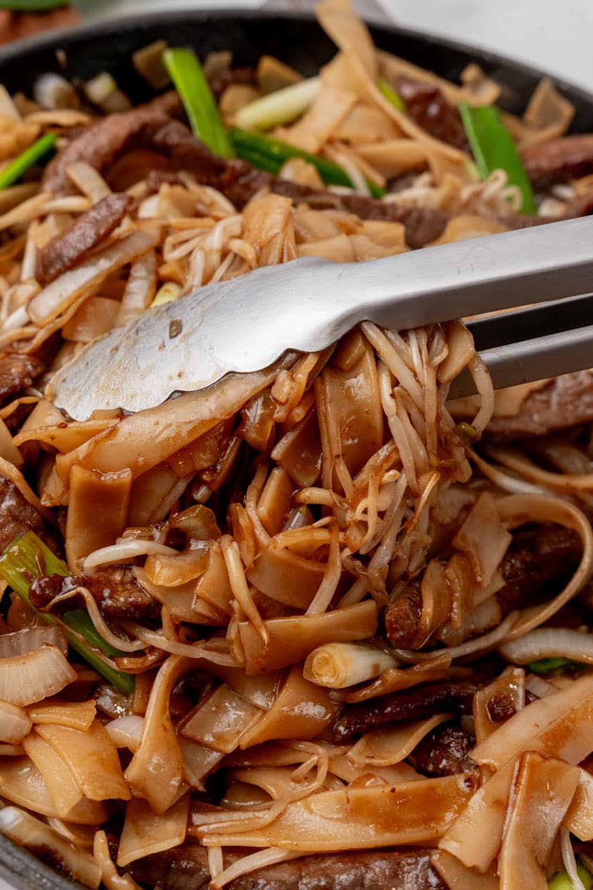 silver tongs scooping beef chow fun out of a large cast iron skillet