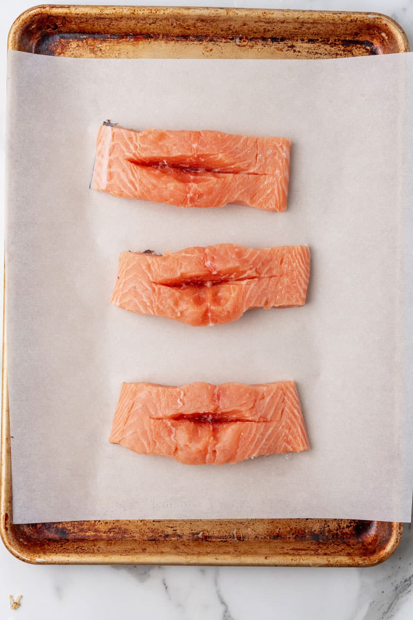 three salmon filets with slits cut in the top arranged in a row on a parchment paper lined baking sheet