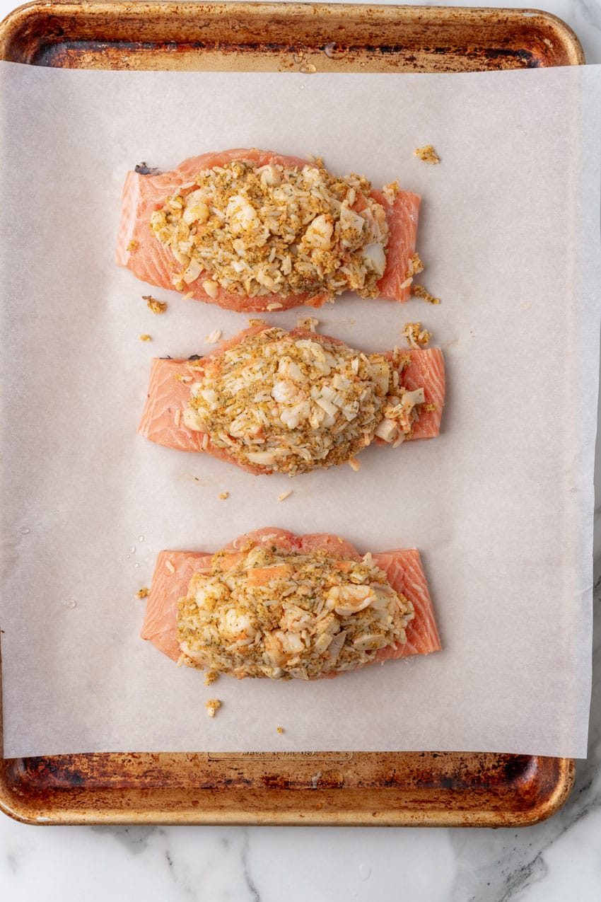 three crab stuffed salmon filets arranged in a row on a parchment paper lined baking sheet