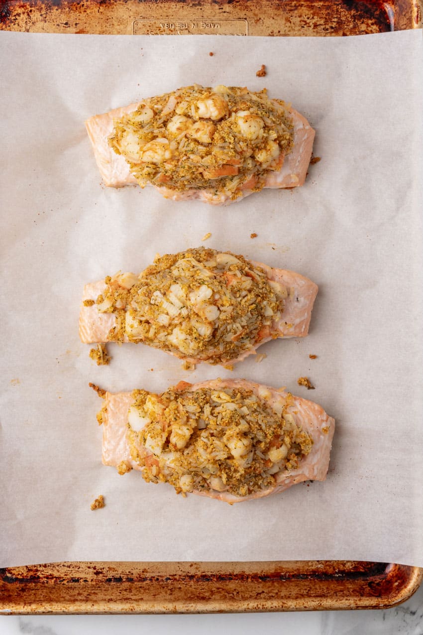three baked crab stuffed salmon filets arranged in a row on a parchment paper lined baking sheet
