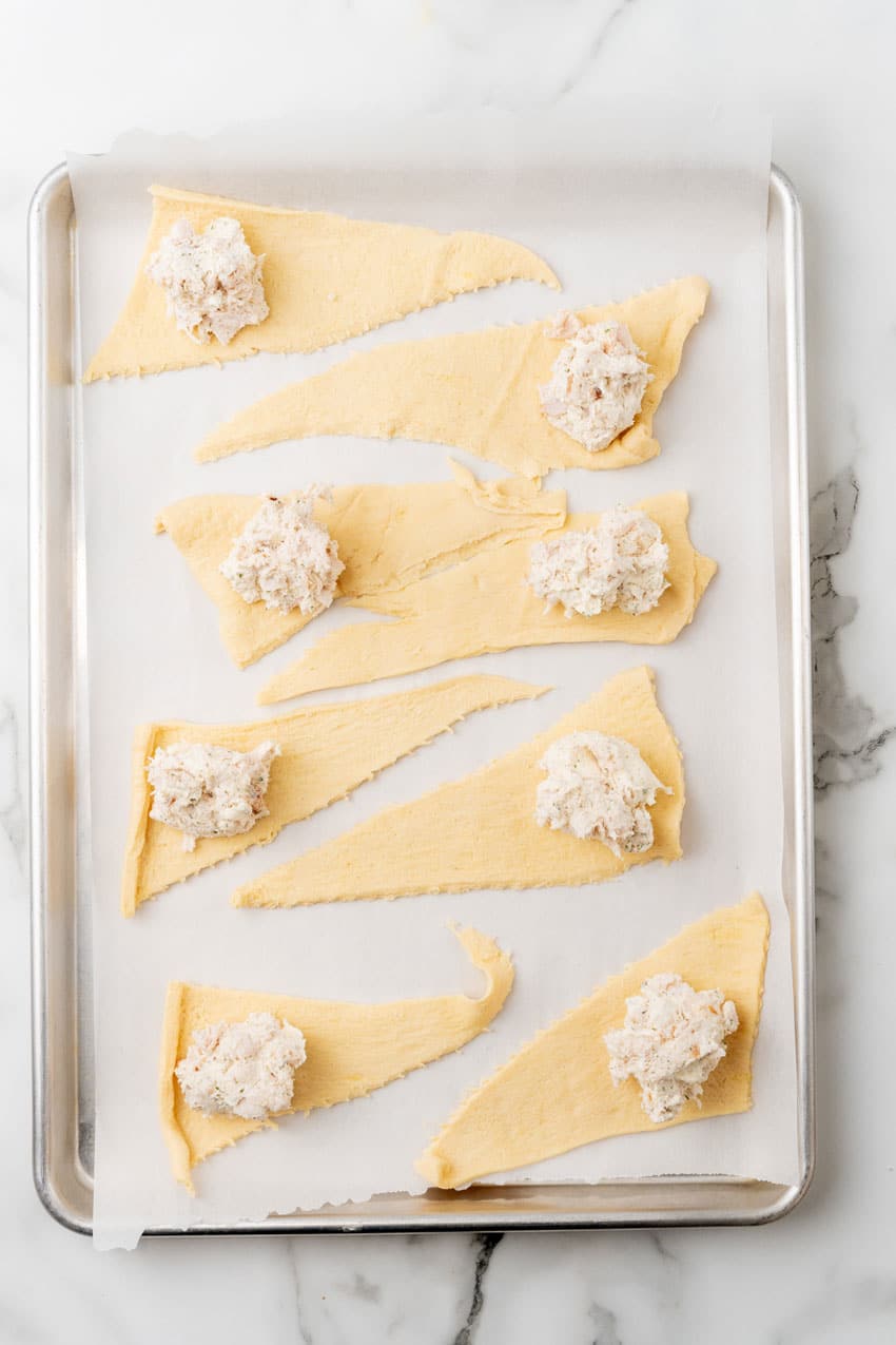 crescent roll dough topped with creamy chicken mixture arranged in a row on a parchment paper lined baking sheet