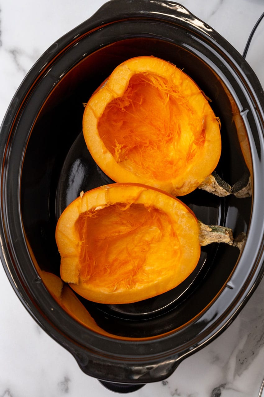 two hollowed out halves of a pie pumpkin in the black bowl of a slow cooker