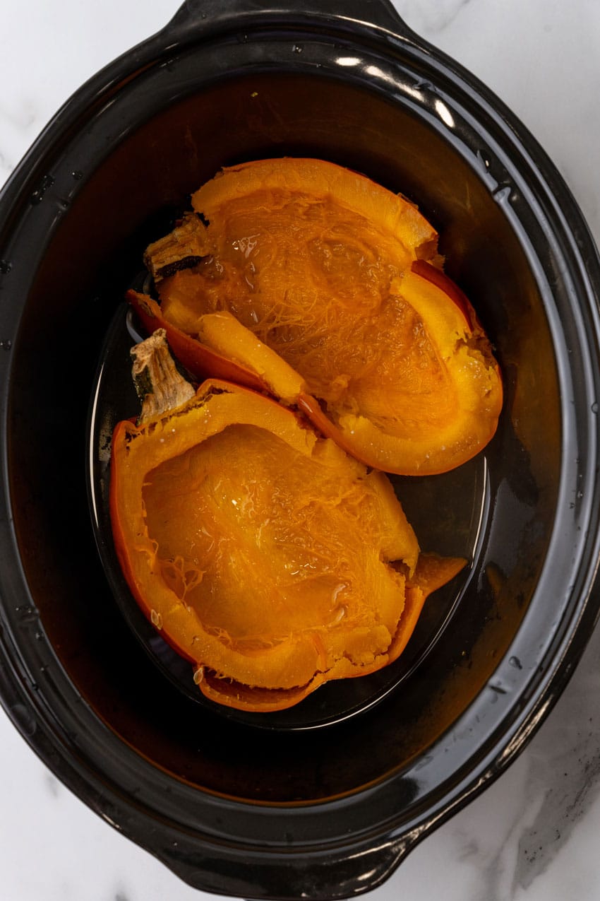 two cooked halves of a pie pumpkin in the black bowl of a slow cooker