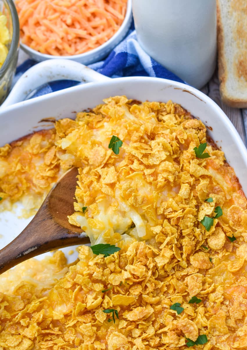 a wooden spoon scooping cheesy hashbrown casserole out of a white baking dish
