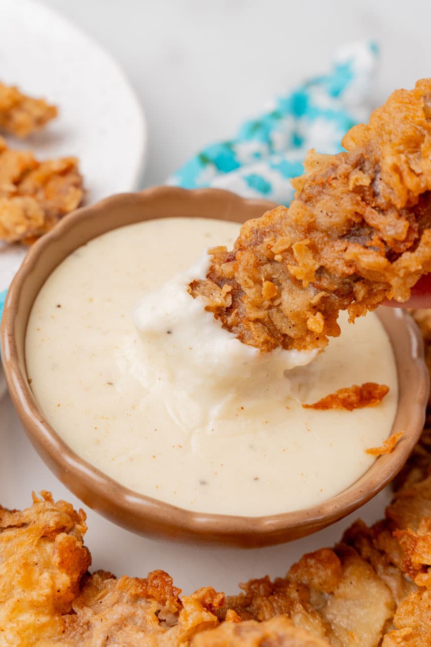 a chicken fried steak fiinger being dipped in a small bowl of homemade creamy white gravy