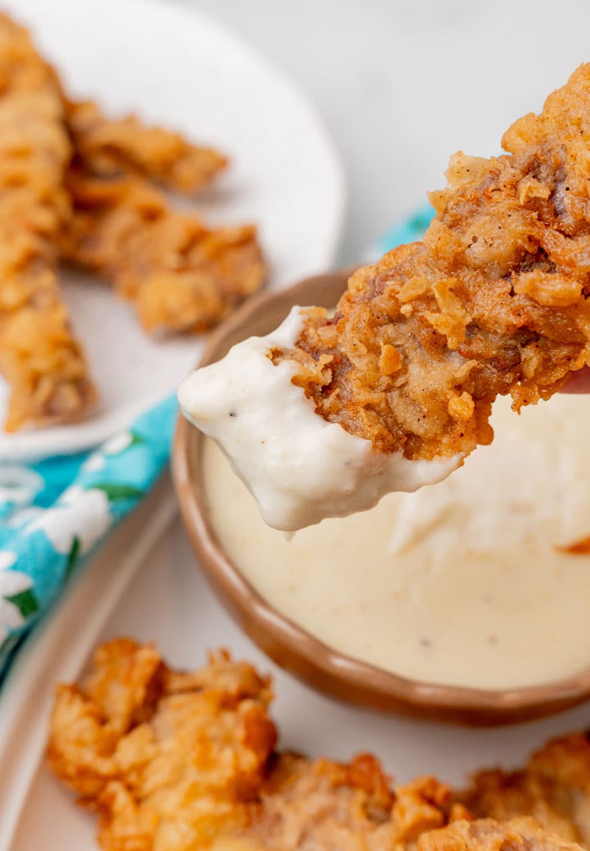 a chicken fried steak fiinger being dipped in a small bowl of homemade creamy white gravy