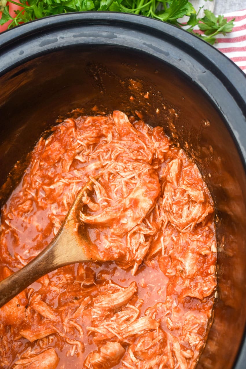 a wooden spoon scooping shredded parmesan chicken out of a black crockpot