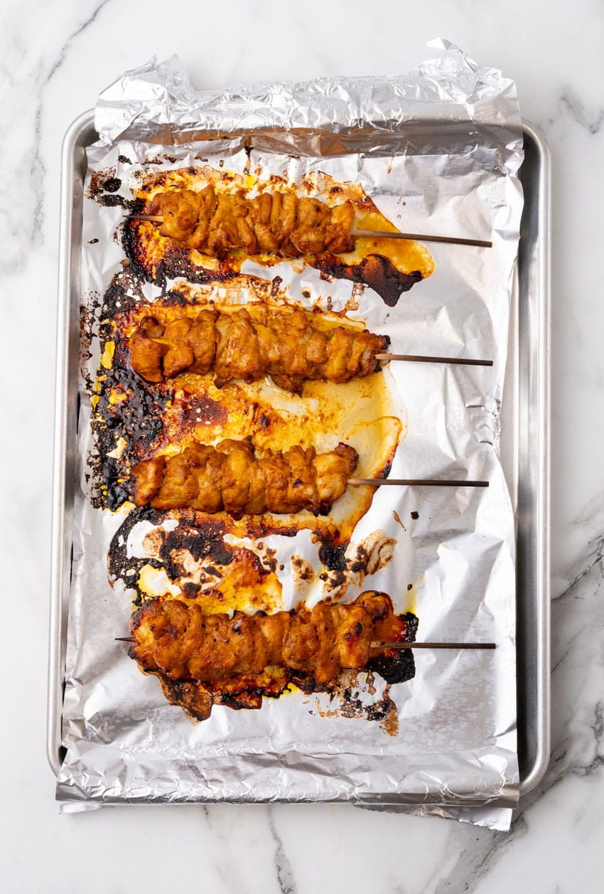 baked chicken satay skewers arranged in a row on an aluminum foil lined baking sheet