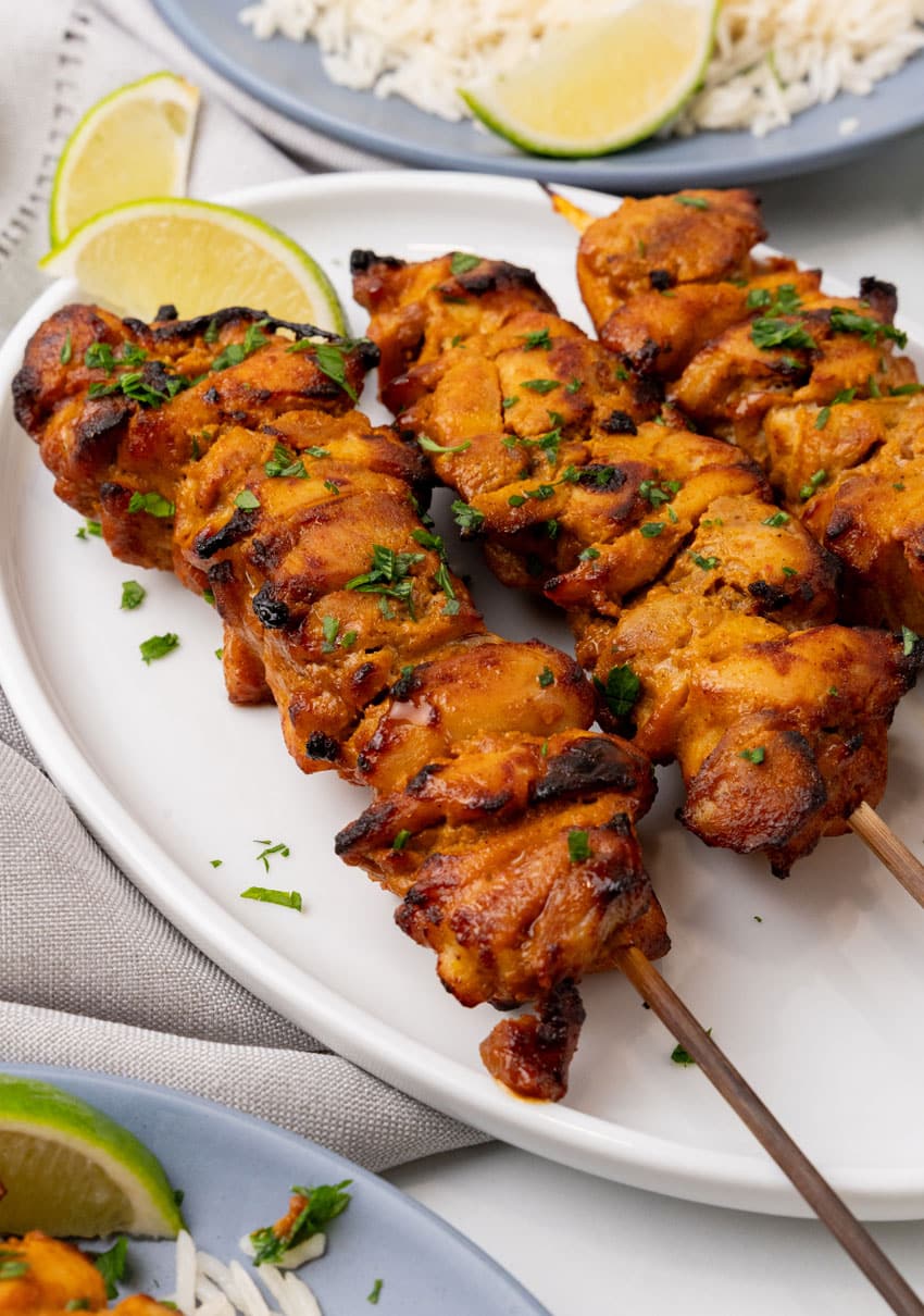 three chicken satay skewers on a white serving plate with fresh cut lime wedges on the side